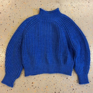 Blå stickad polotröja - Blå stickad polotröja med grov ribbstickning och hög krage. Tröjan har långärmade ärmar med breda muddar och en loose fit som ger en avslappnad vibe. Perfekt för kyliga dagar när du vill vara både varm och stilren.