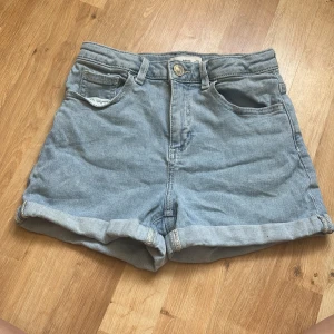 Ljusblå jeansshorts från MNG Denim - Snygga ljusblå jeansshorts från MNG Denim med klassisk femficksdesign och uppvikta benslut. Shortsen har bälteshällor, knapp och dragkedja framtill samt broderad logga bak. Perfekta för varma dagar och enkel att matcha med allt.