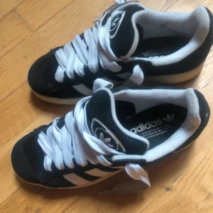 Adidas Campus svarta sneakers mocka - Svarta Adidas Campus sneakers i mocka med klassiska vita ränder och vit sula. Skorna har rund tå, platt gummisula och vita snören. Perfekta för dig som gillar retrostil och streetwear. Adidas-logga på plösen och sidan.