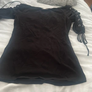 Svart topp med snörning på ärmarna - Säljer en svart topp med cool snörningsdetalj och öljetter på båda ärmarna. Toppen har en tight passform och i bomull. 