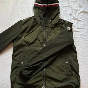 Grön vindjacka från Moncler - Moncler Windbreaker  - Snygg mörkgrön vindjacka från Moncler med huva och dragsko, dragkedja framtill och två fickor med tryckknappar. Scanen funkar se i bild 3. Priset går att diskutera vid snabb affär. (Jackan är i bra skick)