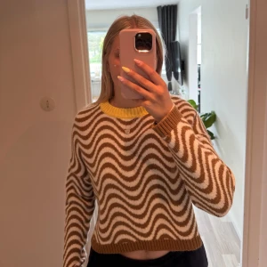 Brunvitrandig stickad tröja Design by Si - Säljer en stickad tröja från Design by Si med vågiga bruna och vita ränder och en ribbad gul krage. Tröjan har lång ärm och är i en skön mix av ull, alpacka och syntet. 