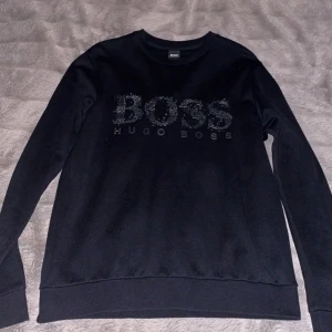 Svart sweatshirt från Hugo Boss - Svart sweatshirt från Hugo Boss med stor BOSS-logga framtill i glansiga svarta detaljer. Tröjan har rund halsringning, långa ärmar och ribbade muddar. Perfekt för en clean och stilren look.