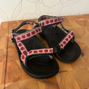Teva sandaler med jordgubbsmönster - Säljer ett par svarta Teva sandaler med sött jordgubbsmönster. Remmarna är justerbara med kardborre. Jättefint skick!!