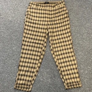 Chinos byxor från Vero Moda L/30 - Snygga rutiga byxor från Vero Moda i beige och svart. Byxorna har elastisk midja och raka ben med avslappnad passform. Perfekta för en chill och trendig look. Mjukt material som känns skönt mot huden.