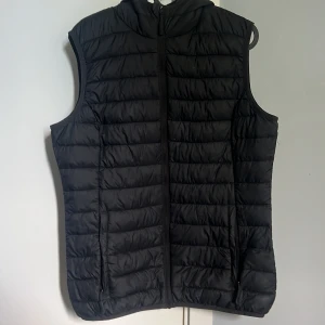 Svart dunväst med huva från Rosetti - Svart quiltad dunväst från Rosetti med huva och hel dragkedja framtill. Västen har två fickor med dragkedja och en stilren, sportig look. Perfekt för lager-på-lager och enkel att matcha med andra plagg.