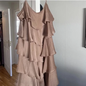 Beige volangklänning med smala axelband - Supersöt beige långklänning med flera lager volanger och smala axelband. Klänningen har en flowy och luftig känsla, perfekt för varma dagar. Materialet känns lätt och svalt, och volangerna ger en riktigt trendig look. Kan skicka den idag, då är den framme till midsommar!