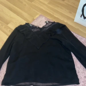 Svart blus med spets och volang - Snygg svart blus med lång ärm och v-ringning. Blusen har spetsdetaljer upptill och en bred volang runt halsen som ger en cool och feminin vibe. Perfekt att styla med jeans eller kjol för en trendig look.