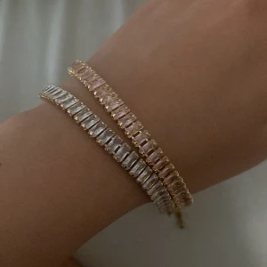 Guldiga armband med stenar - Två snygga armband i guldton med rektangulära stenar, ett med klara stenar och ett med ljusrosa stenar. Perfekta för att ge din outfit lite extra bling och stil. Armbanden har en klassisk och elegant design som verkligen sticker ut. Man kan välja om man vill ha en eller båda en för 30 båda för 50
