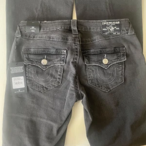 True Religion Joey Low Rise Flare Jeans - Snygga svarta jeans från True Religion, modell Joey Low Rise Flare. Byxorna har låg midja, utsvängda ben och coola ficklock bak med silverfärgade knappar och tydliga kontrastsömmar. Klassisk jeanslook i mörkgrått, perfekt för en trendig streetstil. Byxorna är helt nya!