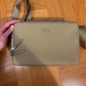 Beige axelväska från Lacoste - Snygg beige axelväska från Lacoste med stilren design och diskret krokodillogga på framsidan. Väskan har justerbar axelrem, dubbla dragkedjor och flera fack inuti med Lacoste-tryck. Perfekt för att bära dina viktigaste grejer med stil.