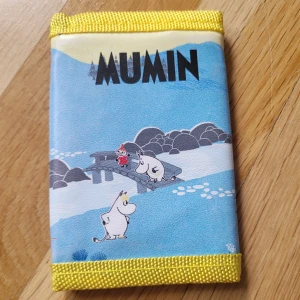 Mumin Moomin wallet - Cute Moomin themed wallet blue with yellow lining. The outside has a printer image of a scene from the Moomin troll world. The front shows Little My/Lilla My, Snorkmaiden/Snorkfröken, and Moomin/Mumin. Little My and Moomin are overlooking the river on a bridge. Moomin is holding a piece of paper. Snorkmaiden is to the side of the river. The back shows Sniff running into trees.  Large zipper pocket, 2 smaller pockets, clear pannel. New, did not come with tags. ©️Moomin Characters ™️ Art nr. 8102048
