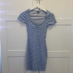 Ljusblå blommig kortklänning H&M Divided - Supersöt ljusblå kortklänning från H&M Divided med små mörkblå blommor, ribbad struktur och lätt rynk vid bysten. Klänningen har korta ärmar och en smickrande v-ringning. Perfekt för sommarens alla tillfällen!