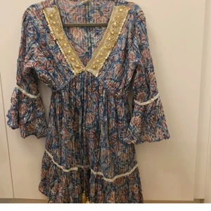 Blommig boho-klänning med volangärm zimmerman inspirerad ONE SIZE - Superfin kortklänning i boho-stil med blått och rosa blommönster, v-ringning med guldiga paljettdetaljer och volangärmar. Klänningen har knytband med tofsar och spetsdetaljer längs ärmar och kjol. Perfekt för dig som gillar färg och mönster!