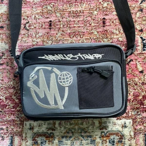 MinusTwo axelväska - Säljer en grå och svart axelväska från MinusTwo med broderad logga och cool graffiti-text framtill. Väskan har justerbar rem, flera fack med dragkedja och en praktisk ficka på framsidan. Perfekt för att bära med sig det viktigaste i vardagen.