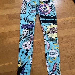 Stronger leggings  - Färgglada leggings från Stronger med pop art-inspirerat tryck i blått, gult, rosa och svart. Text som 'BOOM!' och 'Girl Power!' samt bananer och blixtar. Hög midja med logotyp i midjebandet. Perfekta för dig som vill sticka ut på gymmet.