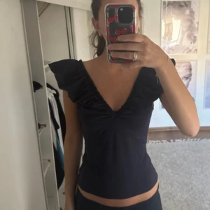 Svart topp med volangärmar och v-ringning - Supersnygg svart topp med djup v-ringning både fram och bak. Toppen har volangärmar som ger en extra touch och är figurnära i modellen. Perfekt att styla med jeans eller kjol för en trendig look. 