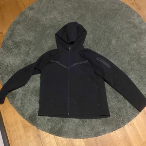 Svart Nike hoodiejacka XL  - Svart hoodiejacka från Nike i storlek XL. Jackan har hel dragkedja, huva och diskreta reflexdetaljer på bröstet och ärmen. Tillverkad i mjukt syntetmaterial, perfekt för träning eller chill. Klassisk Nike-logga på bröstet och sportig look.