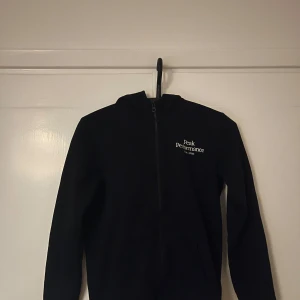 Svart hoodie från Peak Performance - Svart hoodie från Peak Performance med dragkedja framtill och vit logga på bröstet. Tröjan har huva, långa ärmar och två fickor framtill. Perfekt för dig som gillar en clean och sportig stil.