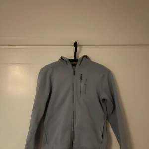 Ljusblå fleecejacka från Sail Racing - Ljusblå fleecejacka från Sail Racing med huva och dragkedja framtill. Jackan har en bröstficka med dragkedja och diskret logga på bröstet samt på huvan. Perfekt för kyliga dagar när du vill vara både bekväm och snygg.