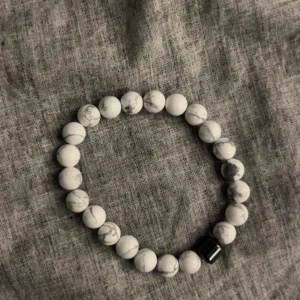Fyra stenkula-armband i olika färger - Säljer fyra snygga armband med runda stenkulor. Ett är vitt med grå marmorering, ett svart med grått mönster, ett brunt med tigeröga-effekt och ett helsvart med lavasten-look. Alla har elastiskt band och en svart cylinderpärla som detalj. Perfekt för dig som gillar stilrena accessoarer.☀️