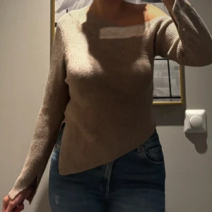 Beige stickad tröja - Säljer en beige stickad tröja med asymmetrisk design och offshoulder-skärning. Tröjan har lång ärm med slits vid ärmslut och en snygg ribbad struktur. 