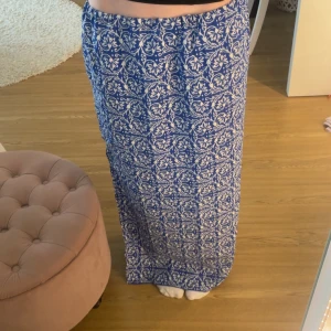 Blåvit långkjol med blommigt mönster - Säljer en flowy blå långkjol med vitt blommigt mönster som jag gjort själv. Kjolen har en rak passform och en snygg slits på sidan som ger extra rörelsefrihet. Perfekt för varma dagar när du vill ha något luftigt och stilrent. Stretchig i midjan