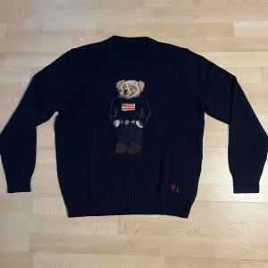 Mörkblå stickad tröja Polo Bear RL - Säljer en ikonisk mörkblå stickad tröja från Ralph Lauren med Polo Bear-motiv framtill. Tröjan har rund hals, långa ärmar och RL-broderi nertill. Perfekt för dig som gillar klassisk preppy stil med en twist. Oanvänd, pris kan diskutera. Lapparna i nacken saknas 
