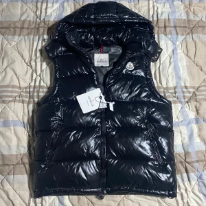 Svart dunväst från Moncler med huva - Säljer en svart, glansig dunväst från Moncler i storlek L. Västen har en avtagbar huva, två dragkedjefickor och klassisk Moncler-logga på bröstet. Perfekt för lager-på-lager och riktigt snygg till streetwear. Stängs med dragkedja framtill. Passar för runt 175-185 cm