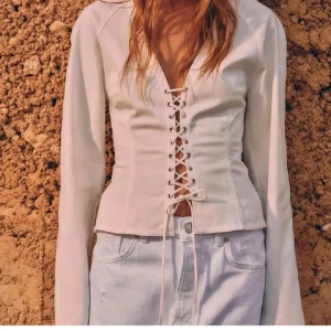 Vit långärmad korsettopp med snörning - Supersnygg vit korsettopp med långa ärmar och djup v-ringning. Toppen har cool snörning framtill som ger en edgy vibe och markerar midjan. Perfekt att styla med jeans eller kjol för en trendig look.