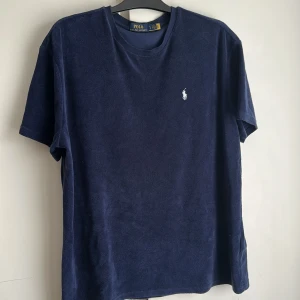 Mörkblå frotté t-shirt Polo Ralph Lauren - Mörkblå t-shirt från Polo Ralph Lauren i mjuk frotté. Klassisk passform med rund halsringning och korta ärmar. Ikonisk vit broderad logga på bröstet. Perfekt för en chill och stilren look.