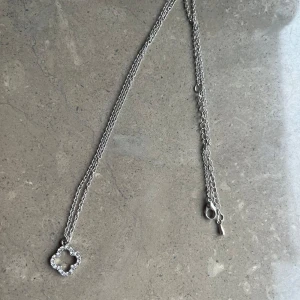 Silverhalsband med glittrig blomma - Snyggt halsband i silverfärg med en tunn kedja och ett hängsmycke i form av en blomma dekorerad med  stenar. Aldrig använt!