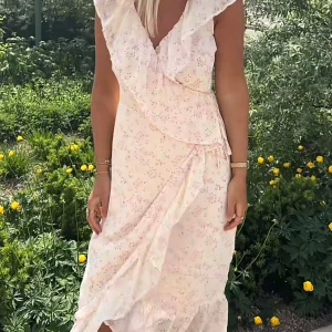 Ljusblommig omlottklänning med volang - Superfin ljus omlottklänning med små blommor i pastellfärger. Klänningen har volangdetaljer längs v-ringningen och kjolen, samt är ärmlös. Perfekt för varma dagar och ger en romantisk vibe.