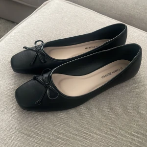 Svarta ballerinaskor Carin Wester 39 - Helt oanvända. Stilrena svarta ballerinaskor från Carin Wester i storlek 39. Skorna har fyrkantig tå, dekorativ rosett framtill och är tillverkade i slätt skinn. Perfekta för en clean och trendig look.