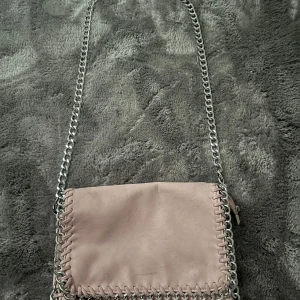 Ljusrosa väska frän Tiamo - Supersnygg ljusrosa axelväska från Tiamo med cool silverfärgad kedja som axelrem och detaljer längs kanterna. Väskan är i mjukt syntetmaterial och har ett stilrent, minimalistiskt uttryck. Perfekt storlek för det viktigaste och ger en trendig touch till din outfit.