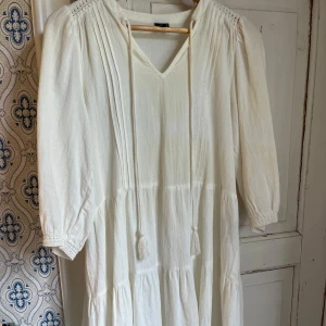 Vit boho-klänning med knytband - Luftig vit klänning i boho-stil med v-ringning och knytband med tofsar framtill. Klänningen har trekvartsärmar med lätt puff och smock vid ärmslut, samt volanger i kjolen för extra flow. Perfekt för varma dagar och en chill vibe.