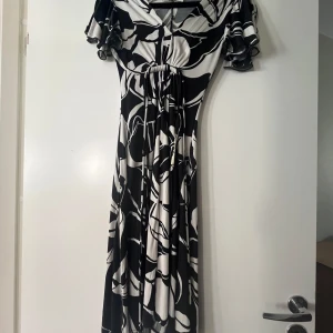 Svart och vit klänning M by Madonna - Snygg långklänning från M by Madonna i svartvitt abstrakt mönster. Klänningen har v-ringning, korta volangärmar och markerad midja med knyt. Mjuk och följsam känsla, perfekt för dig som gillar statement-plagg.
