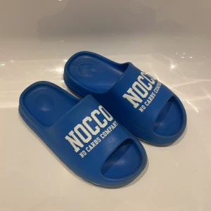 Blåa NOCCO badsandaler stl 38 - Blåa badsandaler från NOCCO med vit text framtill. Skorna har öppen tå och ribbad sula för extra grepp. Tillverkade i syntetmaterial, perfekta för stranden eller poolen.