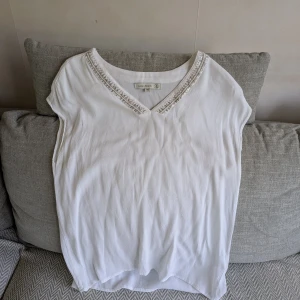 Vit blus med pärldetaljer från Flash Woman - Vit blus från Flash Woman med v-ringning och pärldetaljer längs halsringningen. Jag är ca 170 cm för referens. Vid köp av flera klädesplagg av mig samfraktar jag såklart!