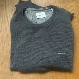 Mörkgrå stickad tröja från GANT - Snygg mörkgrå stickad tröja från GANT med diskret logga broderad på bröstet. Skicket är 9/10 och är bara använd ett fåtal gånger!