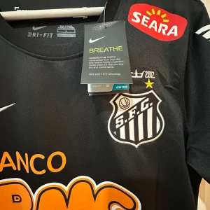 Santos Neymar Jr svart matchtröja Nike - Santos FC fotbollströja med Neymar Jr och nummer 11 på ryggen. Svart med vita och orange tryck, klubbmärke på bröstet och flera sponsorer. Tillverkad i lätt Dri-Fit material som andas. Klassisk rund hals och korta ärmar.