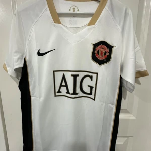 Manchester United vit bortatröja Nike - Manchester Uniteds bortatröja i vitt med svarta och guldiga detaljer. Klassisk klubbmärke på bröstet, svart Nike-logga och AIG som sponsor. Tillverkad i lätt och ventilerande material med Dri-FIT-teknologi. V-ringad krage och korta ärmar.