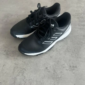Oanvända svarta Adidas golfskor med dobbar - Svarta golfskor från Adidas med vita detaljer och klassiska tre ränder på sidan. Skorna har snörning och en robust sula med dobbar för extra grepp på golfbanan. storlek 38 och säljs för ca 799 nypris.