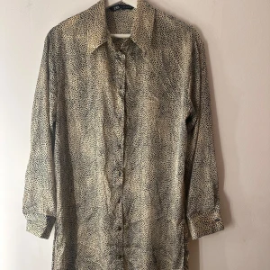 Leopardmönstrad skjortklänning från Zara - Snygg skjortklänning från Zara i beige med svart leopardmönster. Klänningen har knappar hela vägen framtill, klassisk krage och långa ärmar. Perfekt för dig som vill sticka ut med ett coolt mönster och avslappnad passform. Storlek M
