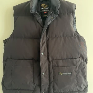 Svart dunväst stl M från Canada Goose - Mkt fint skick sparsamt använd. Svart dunväst från Canada Goose med hög krage och två stora fickor framtill. Västen har tryckknappar och dragkedja, samt diskret logga på ena fickan. 