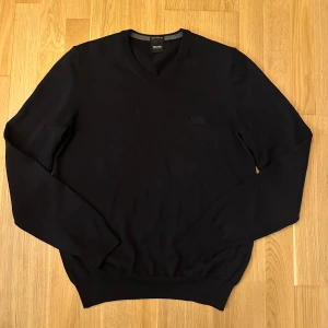 Svart stickad tröja från Hugo Boss - Klassisk svart stickad tröja från Hugo Boss med v-ringning och diskret logga på bröstet. Tillverkad av fin italiensk ullblandning, perfekt för lager-på-lager. Långärmad och med ribbade muddar för en snygg passform. Storlek S. Har tyvärr en reva och ett litet märke, se bild 3 och 4