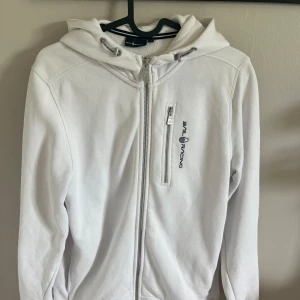 Vit hoodiejacka från Sail Racing - Vit hoodiejacka från Sail Racing med dragkedja framtill och på bröstficka. Broderad logga på bröstet och på huvan. Tröjan är i storlek 170 (tjej). Inköpt för 1000kr men säljer nu för 200kr. Har en liten fläck nedtill (se 4e bild) som inte går bort i tvätten.
