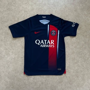 PSG hemmatröja 2023/24 - Paris Saint-Germain fotbollströja från Nike i marinblått med röd vertikal rand och vita detaljer. PSG-logga på bröstet, Qatar Airways-tryck och Nike swoosh i rött. Kortärmad modell i lätt och ventilerande material, perfekt för match eller träning.