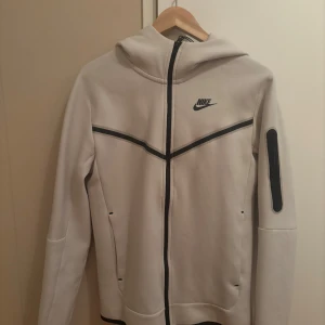 Beige Nike Tech Fleece hoodiejacka - Nike Tech Fleece hoodiejacka i beige med svarta detaljer och dragkedja. Jackan har huva, svarta linjer över bröstet och ärmen samt en praktisk ficka med dragkedja på ärmen. Perfekt för en sportig och clean look. Ordinarie pris 1499 kr enlig unisport.