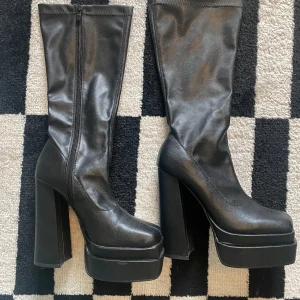 Svarta platåstövlar med hög klack - Snygga svarta stövlar i syntetmaterial med chunky platåsula och hög blockklack. Skorna har fyrkantig tå, dragkedja på insidan och en stilren, minimalistisk look. Perfekta för dig som vill sticka ut med en edgy vibe.